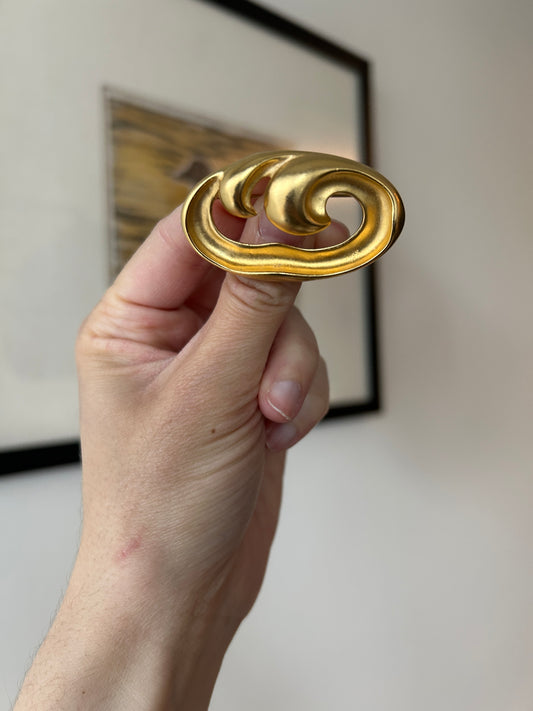 Hand holding a 1980s gold-toned brooch with a shiny, sculptural wave design and an open center.