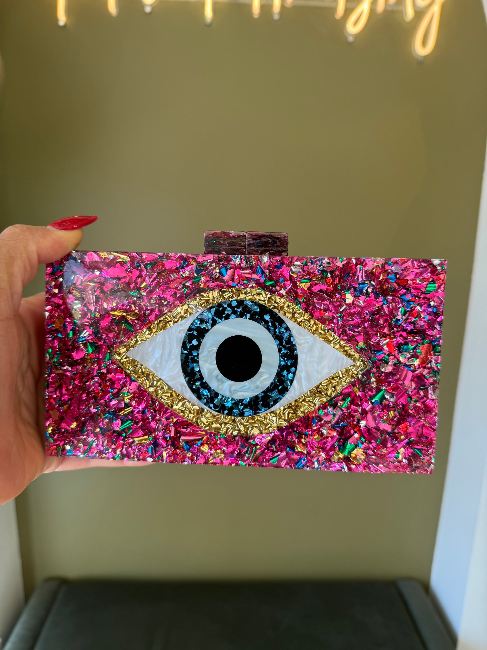Rectangular acrylic clutch featuring a pink confetti design and a central evil eye motif with gold and blue accents.