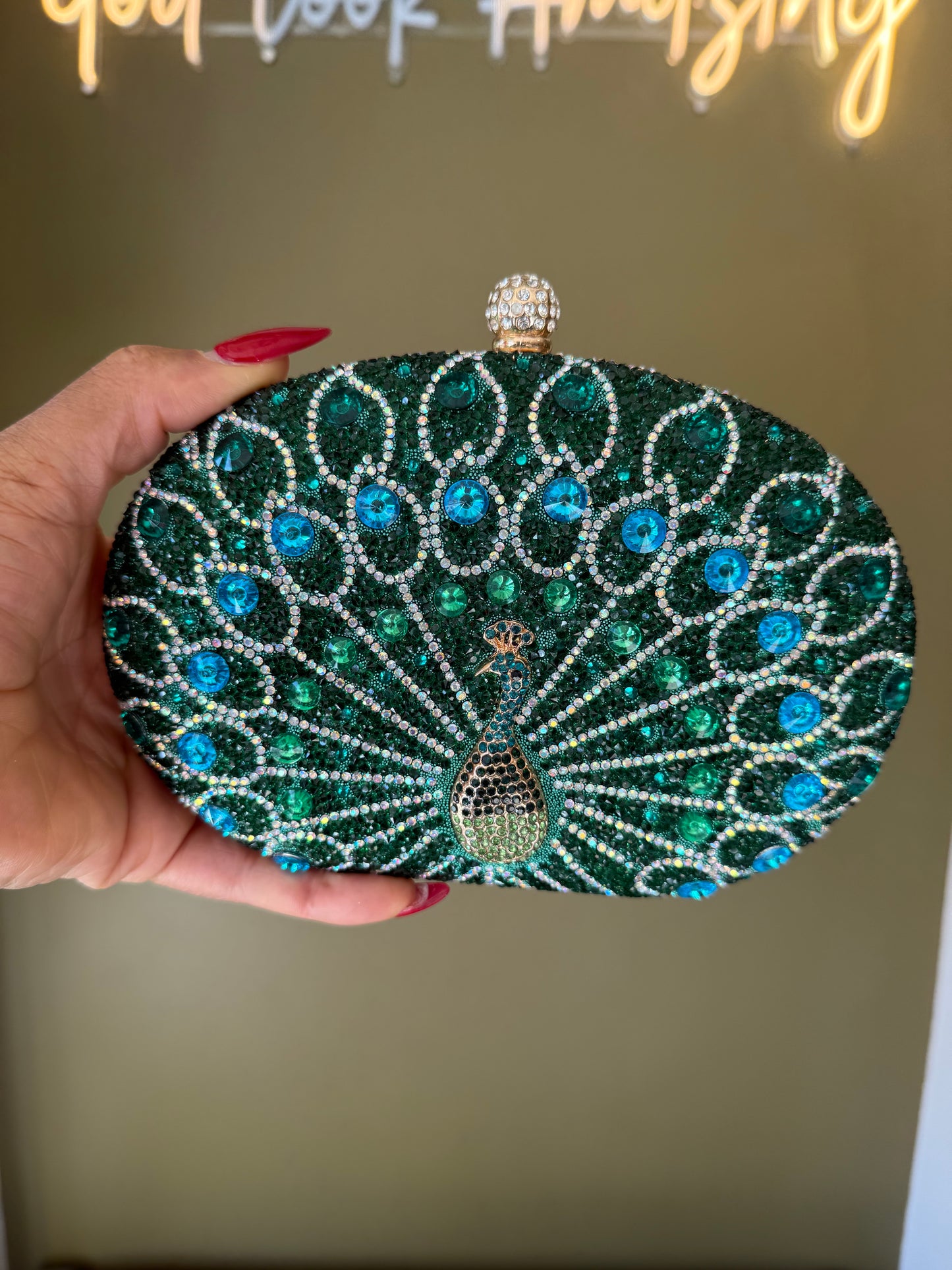 Oval green clutch covered in peacock-patterned rhinestones with a crystal-encrusted clasp held in a hand.