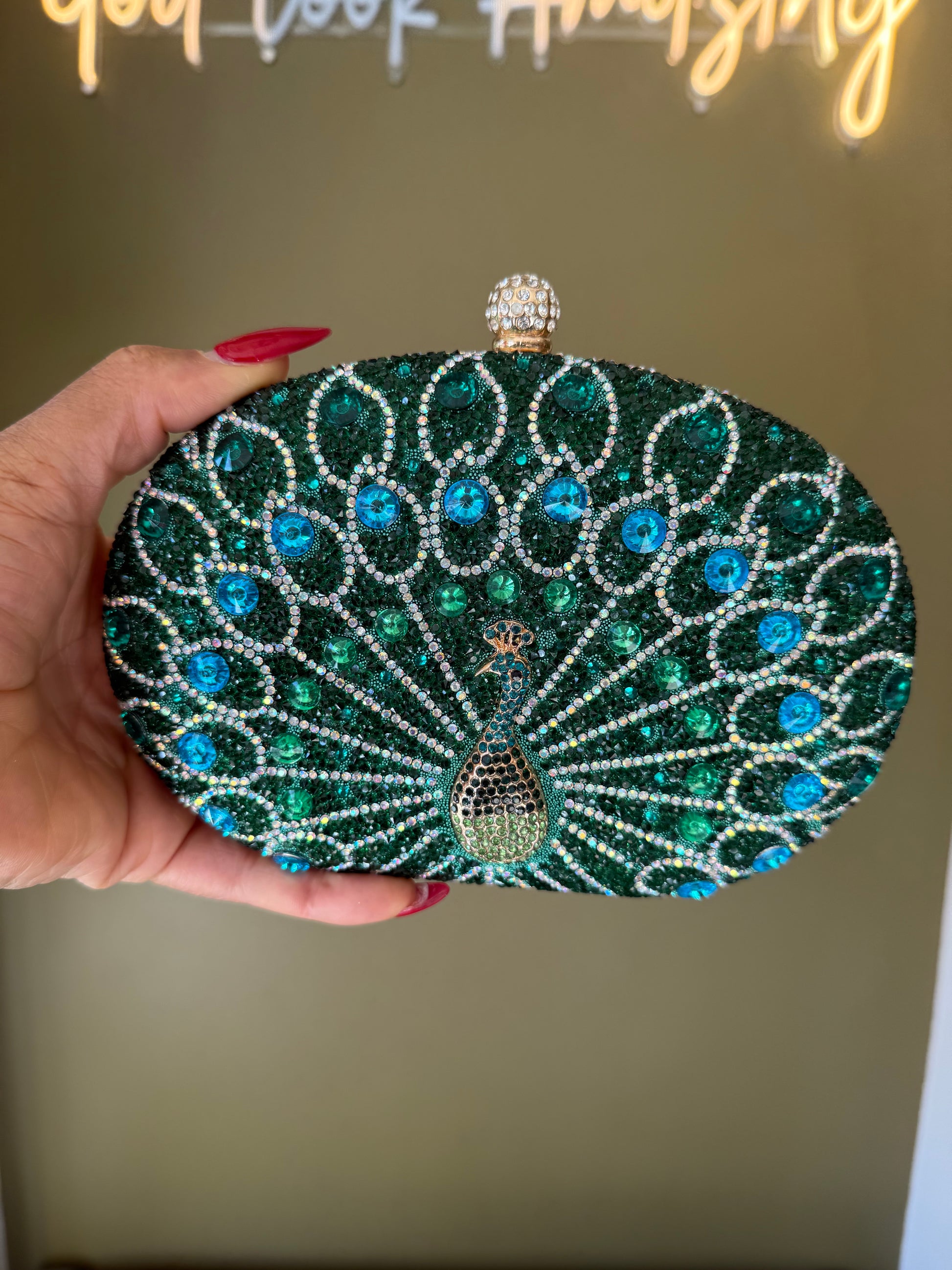Oval green clutch covered in peacock-patterned rhinestones with a crystal-encrusted clasp held in a hand.