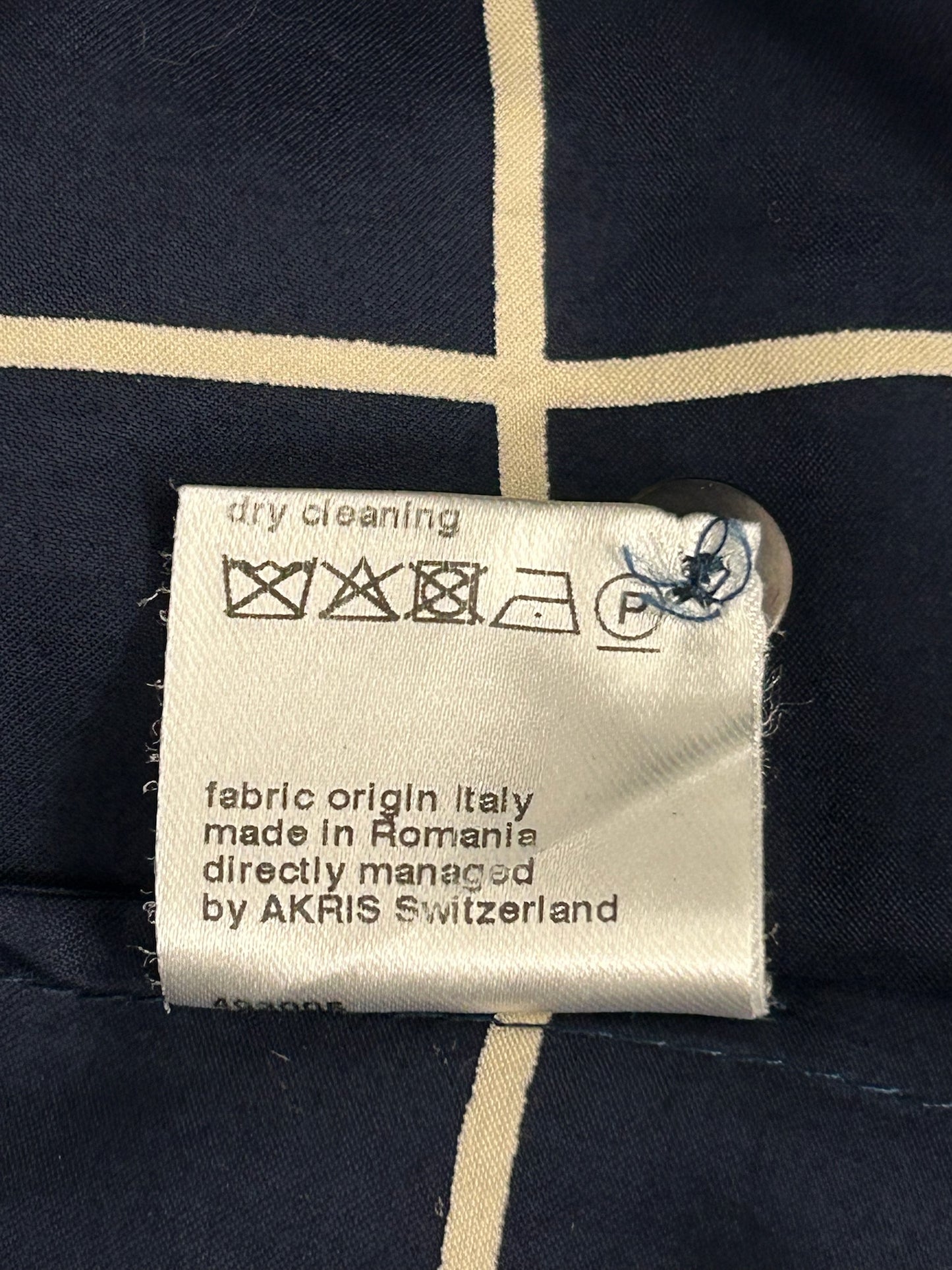 White fabric care tag with dry cleaning symbols, "fabric origin Italy, made in Romania, by AKRIS Switzerland" text, on navy grid pattern.