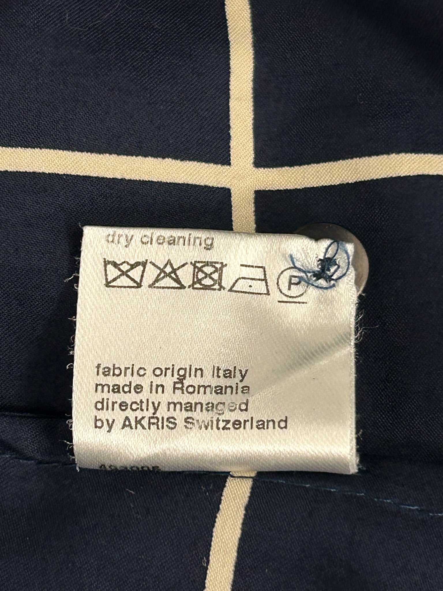 White fabric care tag with dry cleaning symbols, "fabric origin Italy, made in Romania, by AKRIS Switzerland" text, on navy grid pattern.
