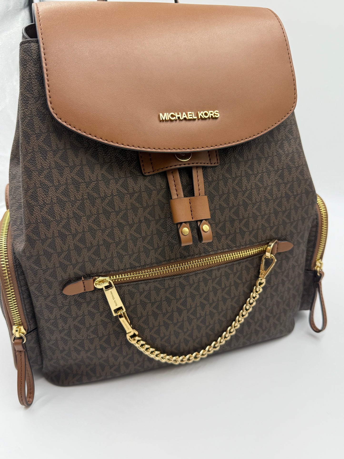 Michael Kors brown monogram Jet Set backpack with tan flap, gold hardware, drawstring, and chain detail.