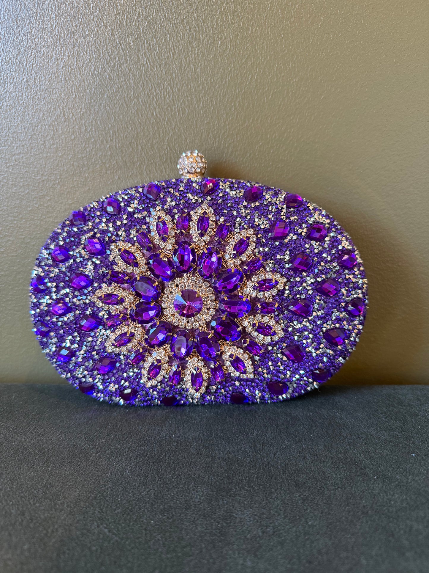 Oval purple clutch covered in sparkling rhinestones with a decorative floral crystal center and gold-tone metal clasp.
