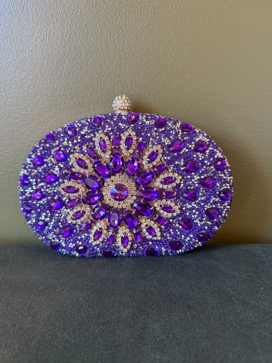 Oval purple clutch covered in sparkling rhinestones with a decorative floral crystal center and gold-tone metal clasp.