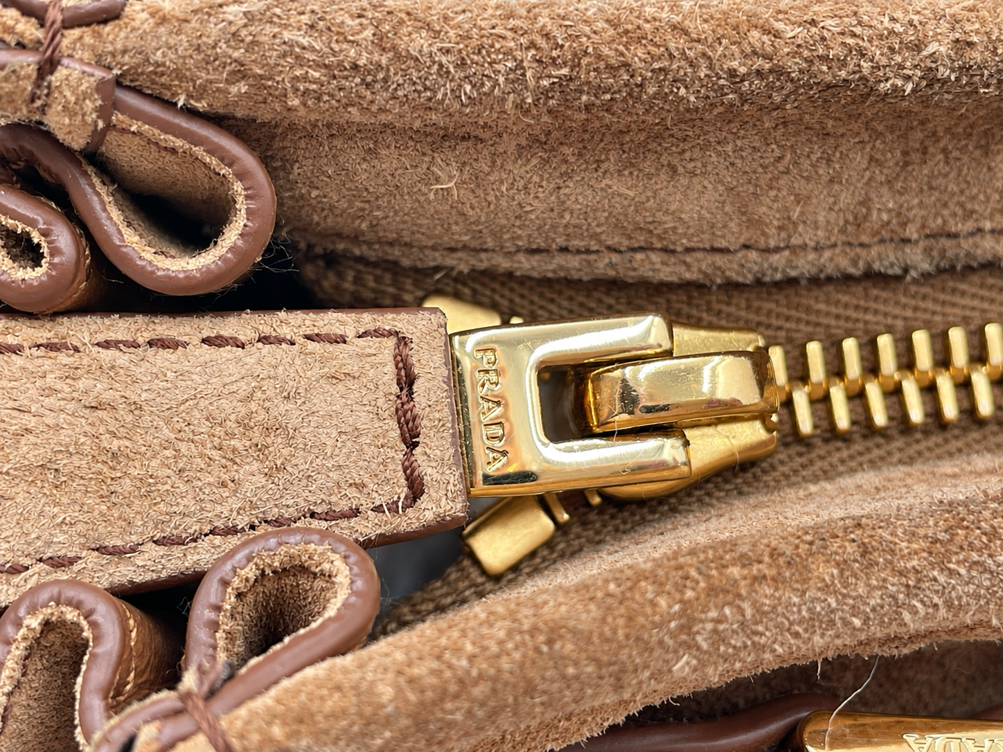 Close-up of a cinnamon brown suede Prada Bonnie bag featuring a gold-toned metal zipper pull with engraved Prada logo.
