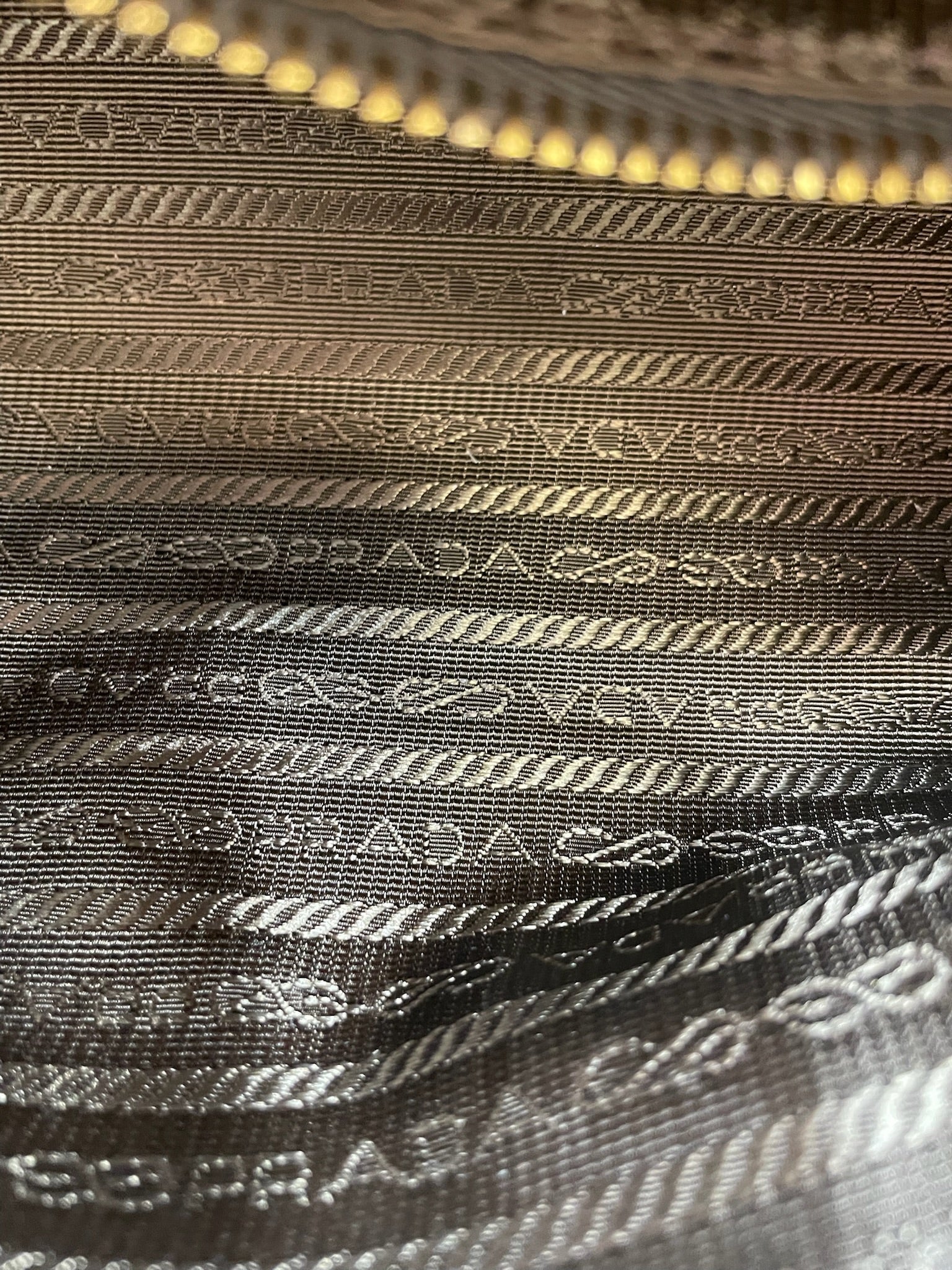 Close-up of Prada Bonnie bag interior featuring brown logo jacquard lining and a gold-tone zipper closure.